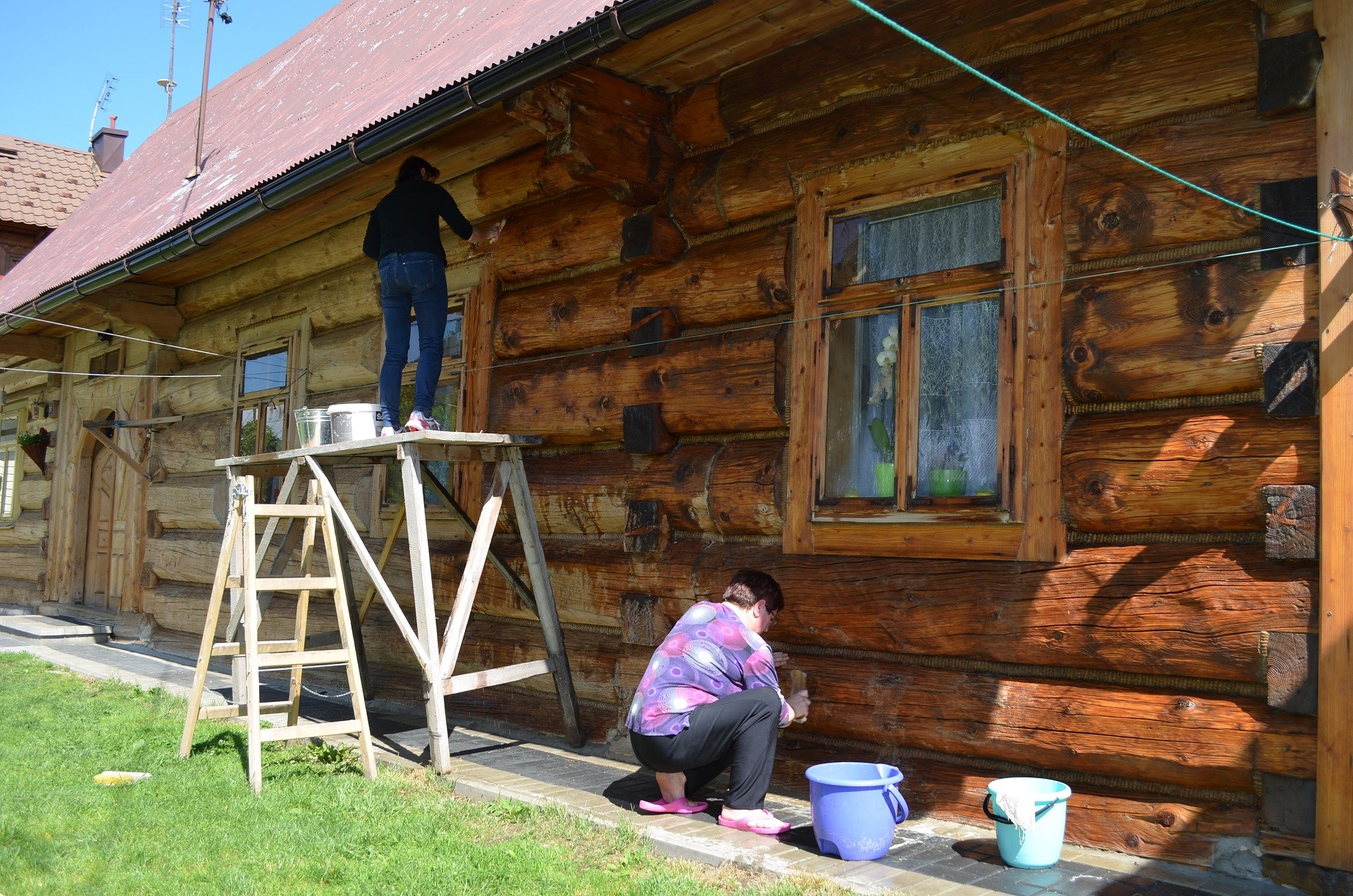 Fragment ściany drewnianego domu, przed nim dwie kobiety, które myją ściany. Jedna stoi na podeście, druga kuca